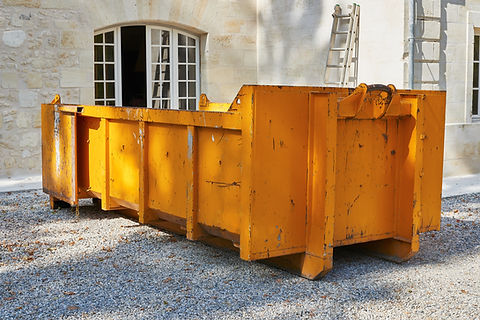 Large orange metal skip sitting on a gravel surface in front of a stone building with arched windows