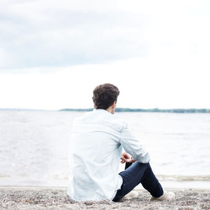 A man in a light shirt sits on a pebbly beach, gazing at a calm, cloudy seascape, creating a serene and reflective mood. This represents a man reflecting on his thoughts, potentially using a thought record.