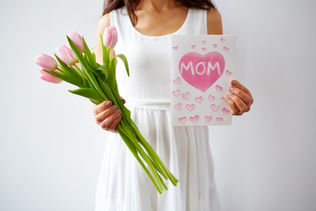 Woman in white dress holds pink tulips and a card with "MOM" in pink heart design. Background is a plain white wall. Mood is loving.