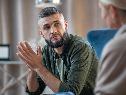 Man in green shirt attentively listening to another person indoors, with a calm, thoughtful expression. Blue chair visible in the background.