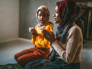 Two Black, femme-presenting people of different ages in hijab are kneeling on a prayer rug with their hands clasped in front of them.