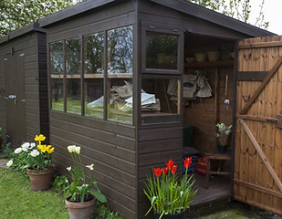Garden shed exterior in Spring, with door open, lawn, tools, flowers, and plant pots
