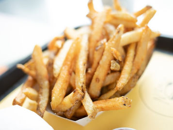 A close-up of golden French fries in a tray with a small cup of ketchup, and part of a burger on a bright table.