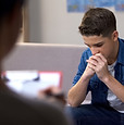 A boy sitting on a couch with his hands folded neatly in his lap, looking contemplative.
