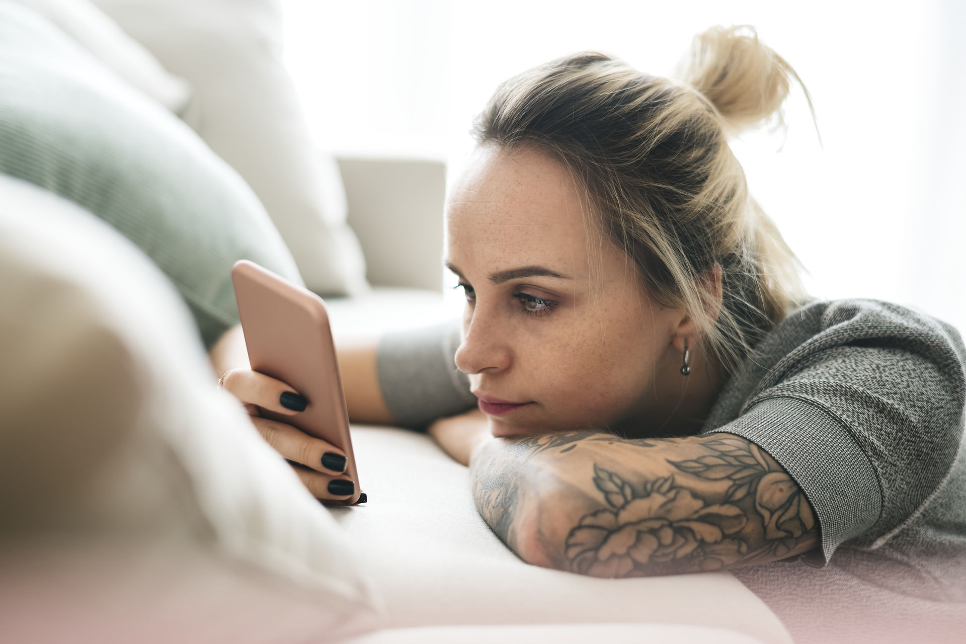 woman with tattoos and blond hair and dark nail polish laying on her bed looking at her phone screen
