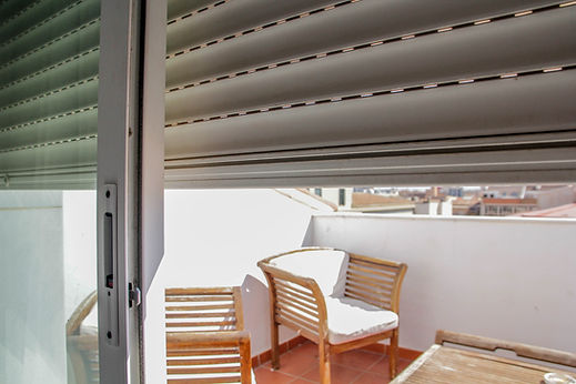 Partially opened white window shutters looking out onto an outdoor balcony area with two wooden chairs