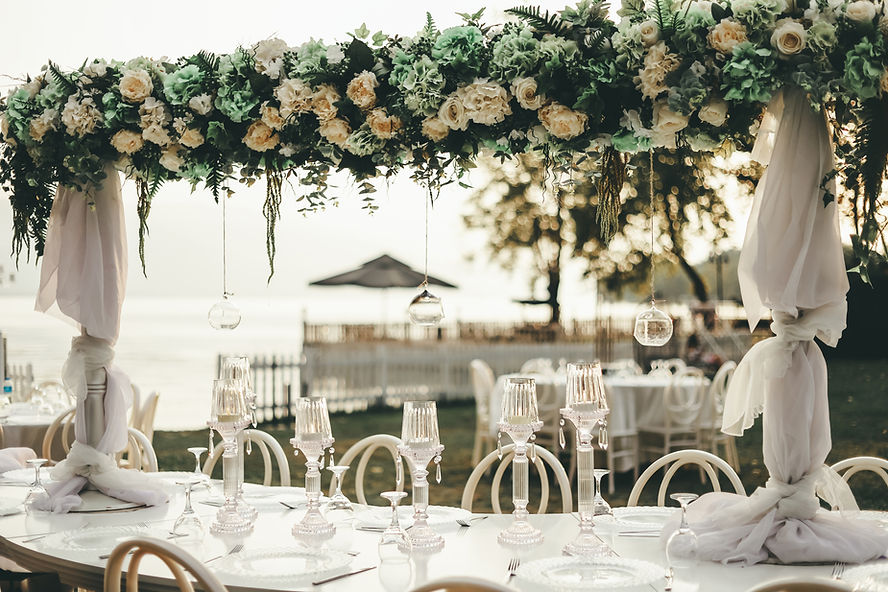 A wedding reception table set with white tablecloths, gold chargers, white dinner plates, gold silverware, two wine glasses, a water glass, and a centerpiece of white flowers and candles.