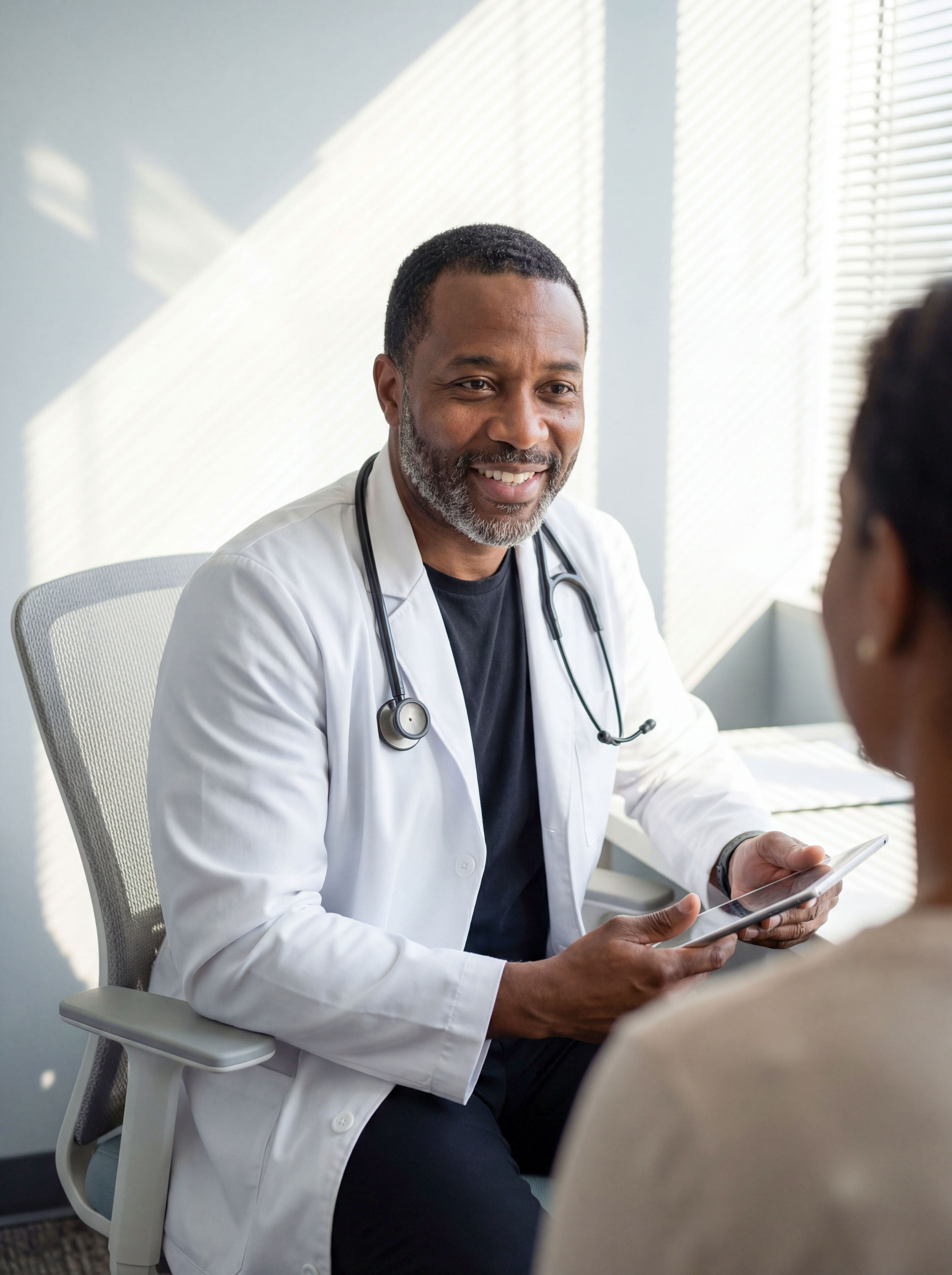 Doctor with a stethoscope holding a tablet and talking to a patient in a well-lit room.