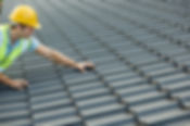 A construction worker wearing a yellow hard hat and safety vest inspects gray roof tiles