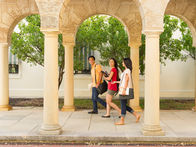 Three smiling students walk under stone arches on a sunny campus, carrying books and bags.