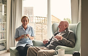 Indoor shot of smiling senior man and female carer enjoying coffee in living room