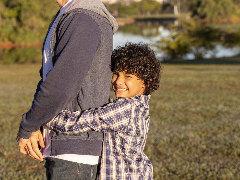 Smiling curly-haired boy hugs an adult from behind on a sunny grassy lakeside, warm and affectionate.