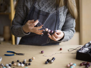 Woman in a gray sweater making bead jewelry at a wooden table with tools and colorful beads scattered around. Creative, focused mood.