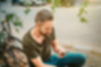A man with a neatly trimmed beard is seated outdoors on a sunny day thinking about making a phone call