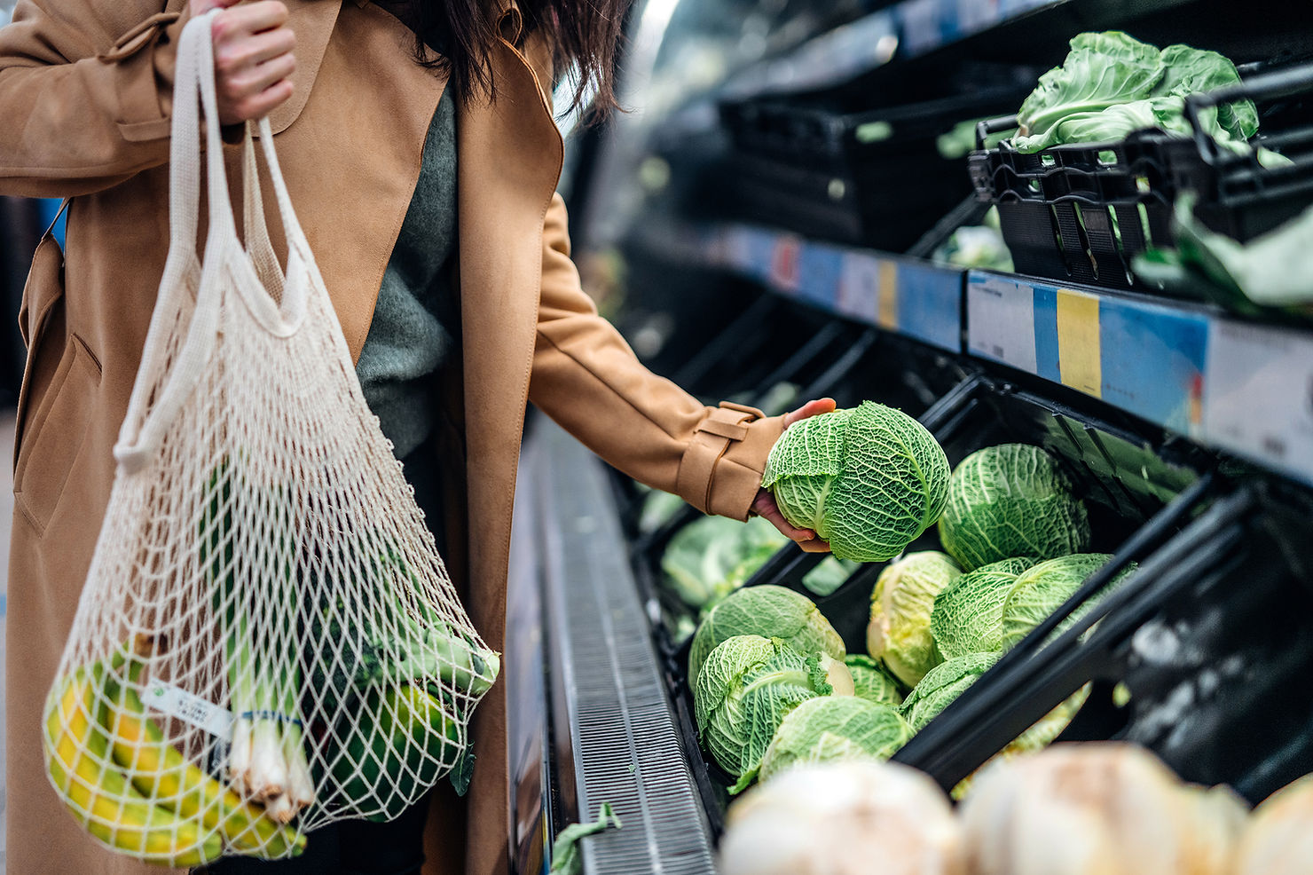 Person in a tan coat shops for cabbages at a grocery store, holding a mesh bag with bananas. Green cabbages line the shelves.
