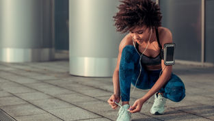 Woman in blue leggings ties shoelaces, wearing earbuds and phone armband, on urban sidewalk with metallic columns in background.