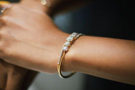 Close-up of a wrist wearing a gold bracelet with sparkling gemstones, against a blurred dark background, highlighting the jewelry's elegance.