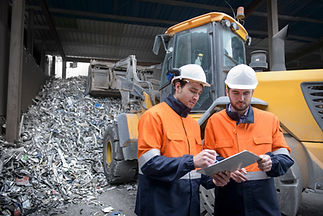 Workers in orange safety gear studying a clipboard near metal scrap and tractor.