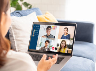 Person on a couch video conferencing with four smiling people on a laptop; bright living room, blue and yellow pillows, relaxed mood.