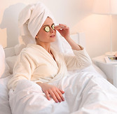 A woman in a white bathrobe and white bath towel on her head with cumbers on her eyes relaxing, lying in bed.
