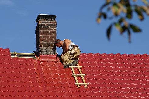 A man wearing a sleeveless shirt and khaki pants works on a brick chimney atop a bright red metal roof