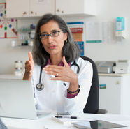 Doctor with gray hair and glasses explains to a patient in an office. She's gesturing with her hands, wearing a white coat, laptop open.