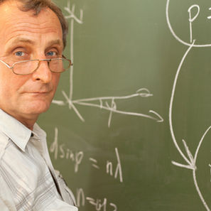 Elderly man with glasses stands by a chalkboard with equations, wearing a light shirt. He looks pensive, background is green.