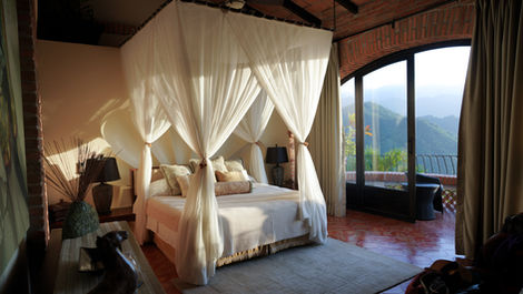 Four-poster bed with white curtains in a sunlit room. Brick walls, terracotta floor, large window view of green hills. Cozy and serene.