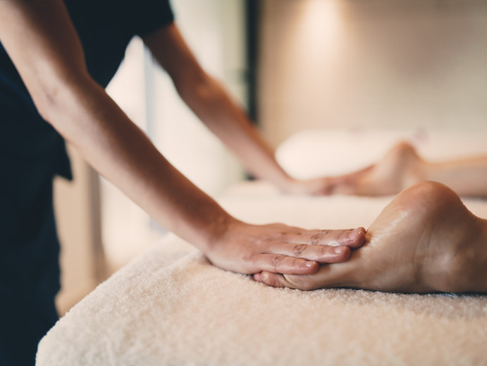 A client is resting face down on a massage therapy table while the practitioner gently holds their feet in a meditative stance. 