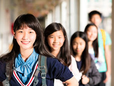 Smiling students with backpacks in a hallway. The front student wears a blue shirt and black sweater. Bright, cheerful setting.