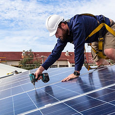 A worker in a white helmet installs solar panels on a roof, using a drill