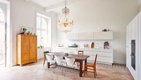 Bright kitchen with a wooden table, mix of white and wooden chairs, chandelier, and wooden cabinet. Large windows and white cabinets.