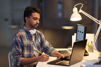 Man in plaid shirt works at a desk with laptop and monitor, wearing headphones around neck. Warm lighting creates a focused mood.