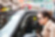A woman in a tan coat and glasses leans toward a black taxi on a city street