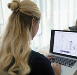 A woman with long blonde hair sits at a desk, using a laptop displaying architectural designs