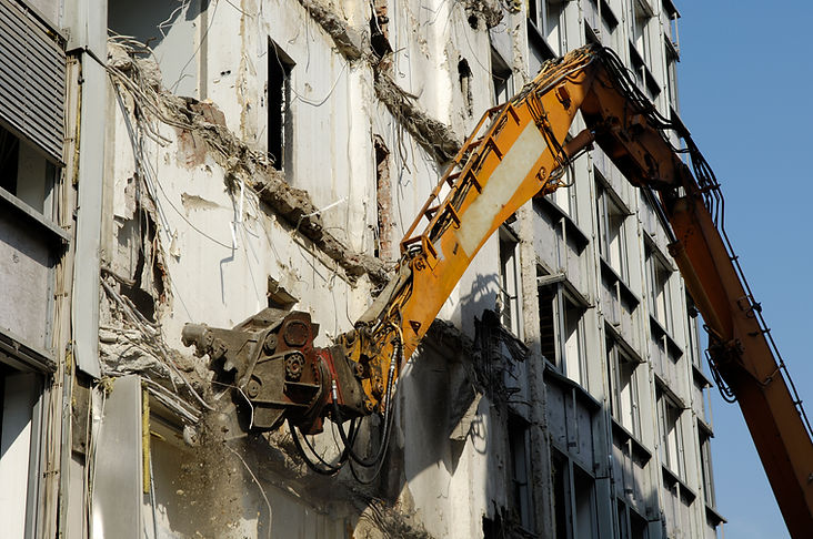 A large excavator arm demolishes an old building, creating debris and dust