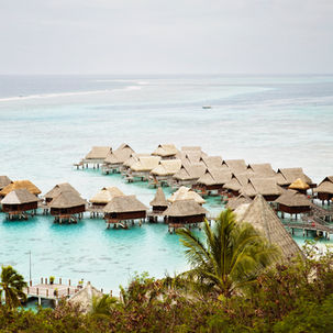 Overwater bungalows with thatched roofs on turquoise lagoon, surrounded by palm trees. Calm ocean in the background under a cloudy sky.
