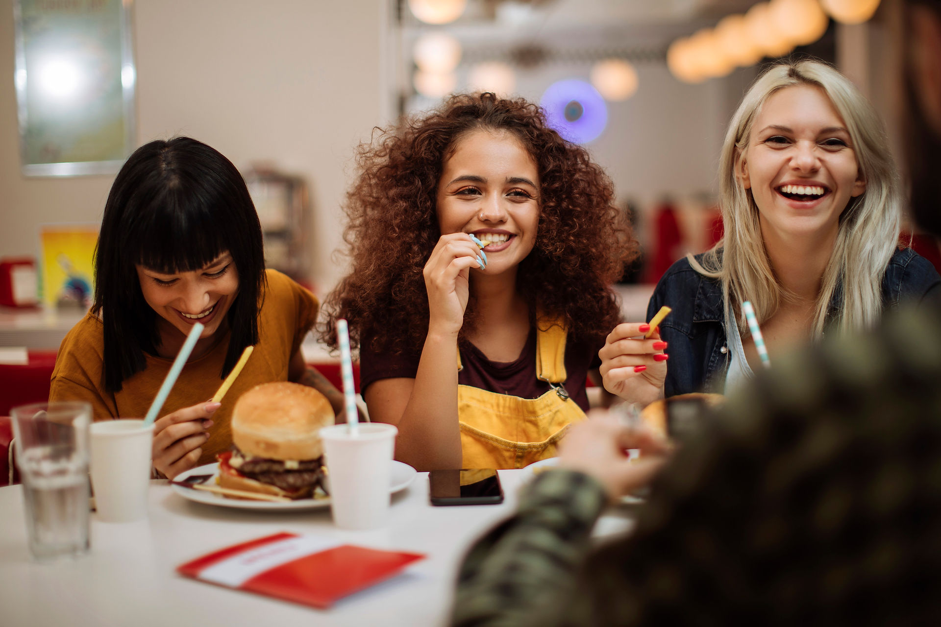 Four young girls of various races and ethnicities sit at a table with burgers and drinks.