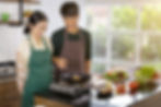 A smiling couple cooks in a bright kitchen. They wear striped aprons and are frying food. Fresh vegetables are on the counter.