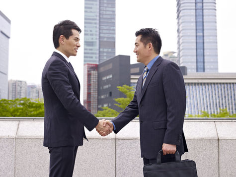Two businessmen in formal attire shake hands outdoors against a backdrop of modern skyscrapers, symbolizing a successful business agreement or partnership.