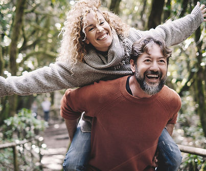 A joyful couple outdoors in a lush forest setting. The man, wearing a rust-colored sweater, is giving the woman, dressed in a cozy gray sweater, a piggyback ride. Both are smiling brightly, and the woman playfully extends her arms like she’s flying.