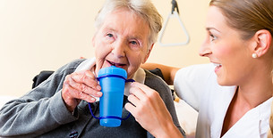 Nurse helping an elderly woman in a wheelchair drink from a blue two-handled cup