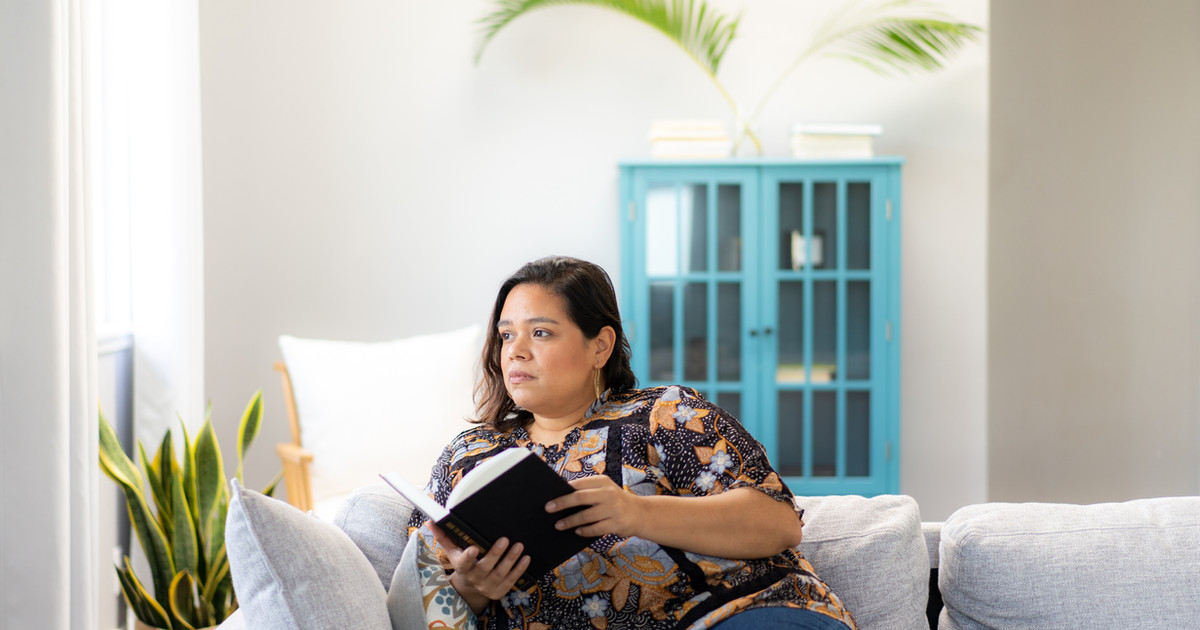 A person sitting on a couch looking thoughtfully out a window, with soft afternoon light, appearing reflective and calm.