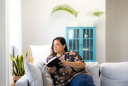 A woman sitting on a comfortable grey sofa, holding an open book and looking thoughtfully off to the side.