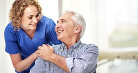 Blonde female carer in blue scrubs talking to a smiling senior man in a striped shirt in a nursing home