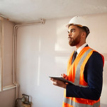 A man in a safety vest holds a clipboard, inspecting an empty room with white walls