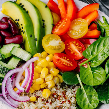 Colourful salad bowl with spinach, tomatoes, corn, avocado, beans, and quinoa. Fresh ingredients arranged in a white dish on a grey table.