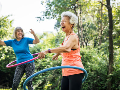 Two women joyfully hula hooping in a sunny park. One wears a blue shirt and leggings, the other in orange. Lush green trees surround them while working on functional balance activities