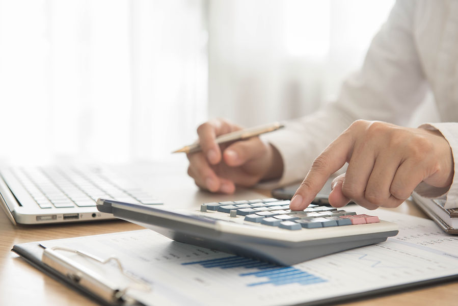 A person uses a calculator at a desk, surrounded by documents, a clipboard, and a laptop