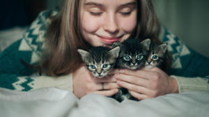 Smiling person holding three kittens on a bed. The person wears a patterned sweater. The mood is cozy and affectionate.